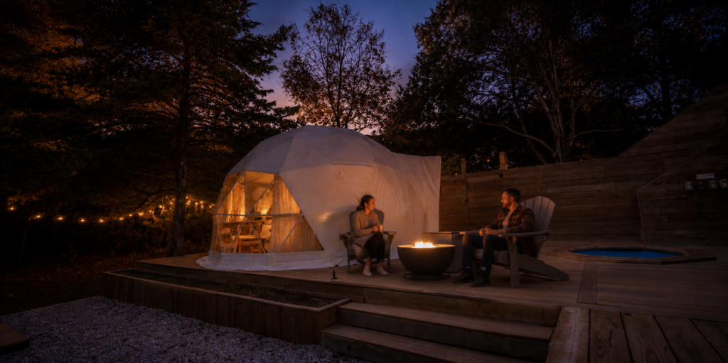 A couple relaxes in separate Adirondack chairs beside a glowing fire pit outside a luxury canvas glamping tent at dusk, surrounded by trees, warm lights, and a private hot tub.